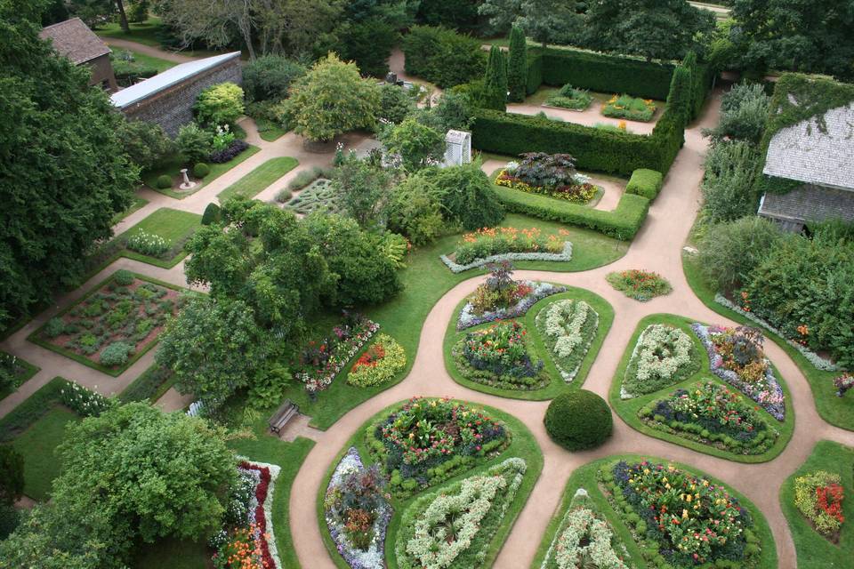 Aerial view of a beautifully landscaped garden featuring colorful flower beds, neatly trimmed hedges, and winding pathways surrounded by trees.