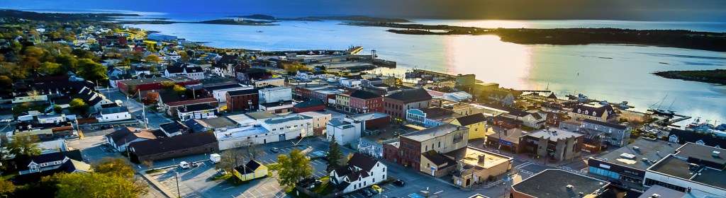 Aerial view of a coastal town with a harbor, featuring a mix of residential and commercial buildings along the waterfront, illuminated by sunset reflections on the water.